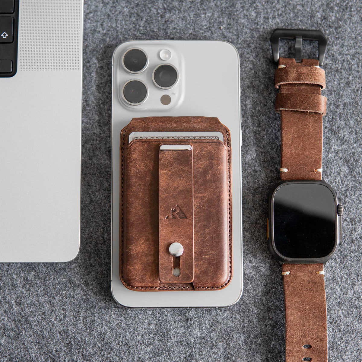 Magnetic cardholder in brown leather attached to a smartphone, with a smartwatch and laptop on a gray surface.
