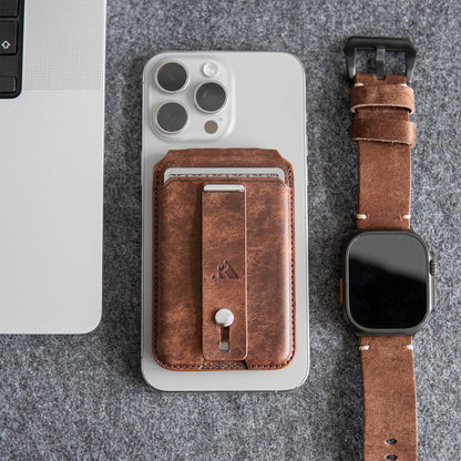 Magnetic cardholder in brown leather attached to a smartphone, with a smartwatch and laptop on a gray surface.
