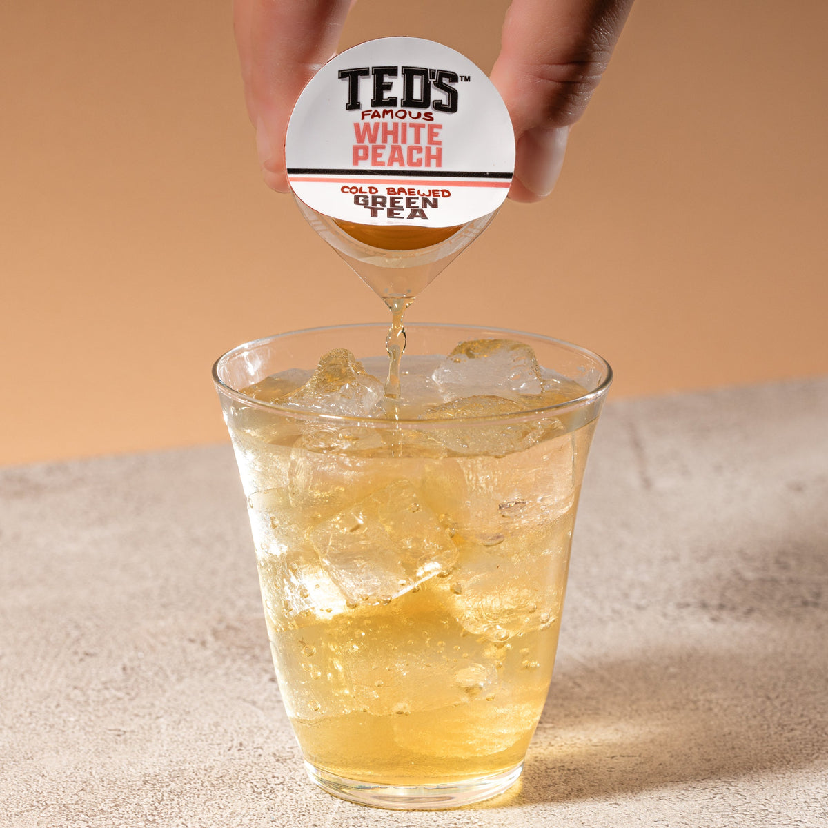 A glass of Ted's Famous White Peach Cold Brew Green Tea by Java House with ice and fresh peach slices on a wooden table.