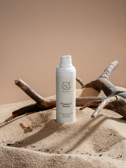 Vitamin C serum bottle from Belleviecosmetic on sand with driftwood, showcasing a 30ml energizing brightening and firming skincare product.